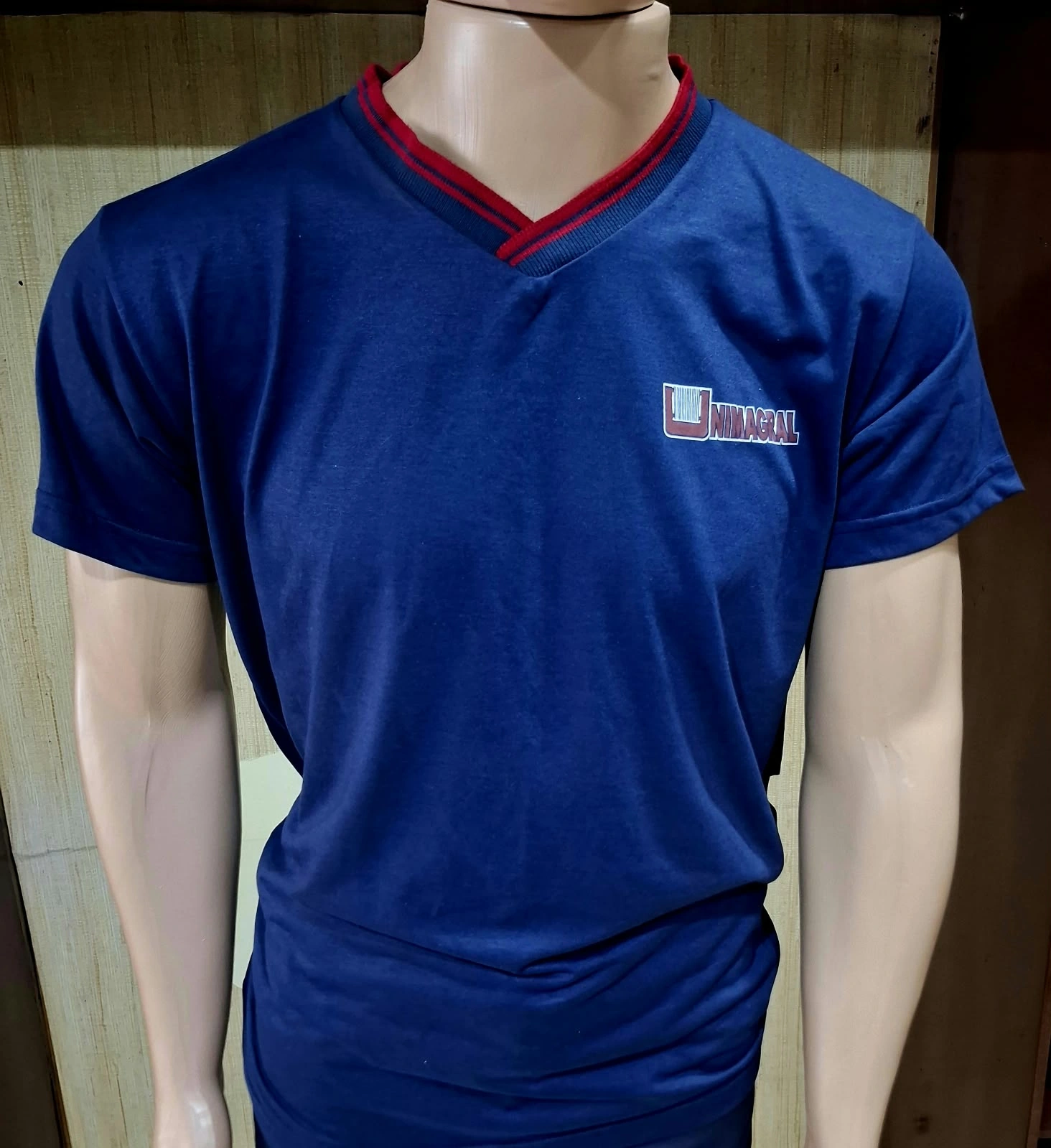 Manequim com camiseta azul, gola em V com detalhe vermelho e logotipo no peito.