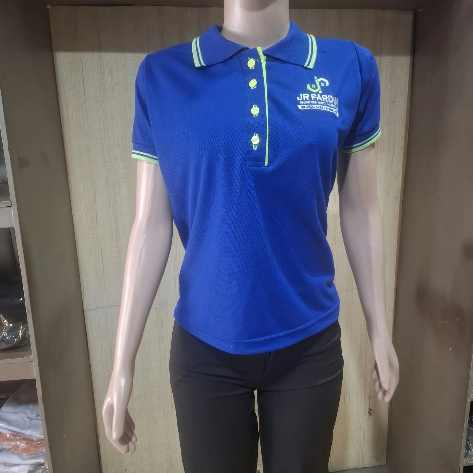Manequim feminino com camisa polo azul, gola e mangas com detalhe verde, botões verdes e logotipo JR Fardas bordado no peito.