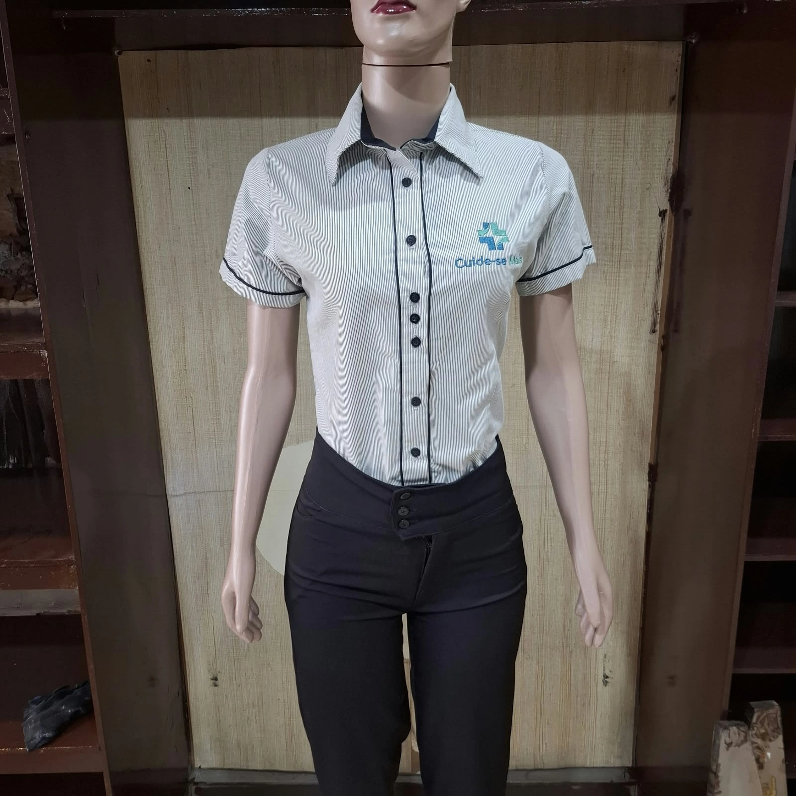 Manequim com camisa cinza de manga curta, botões escuros e bordado no peito, e calça preta.