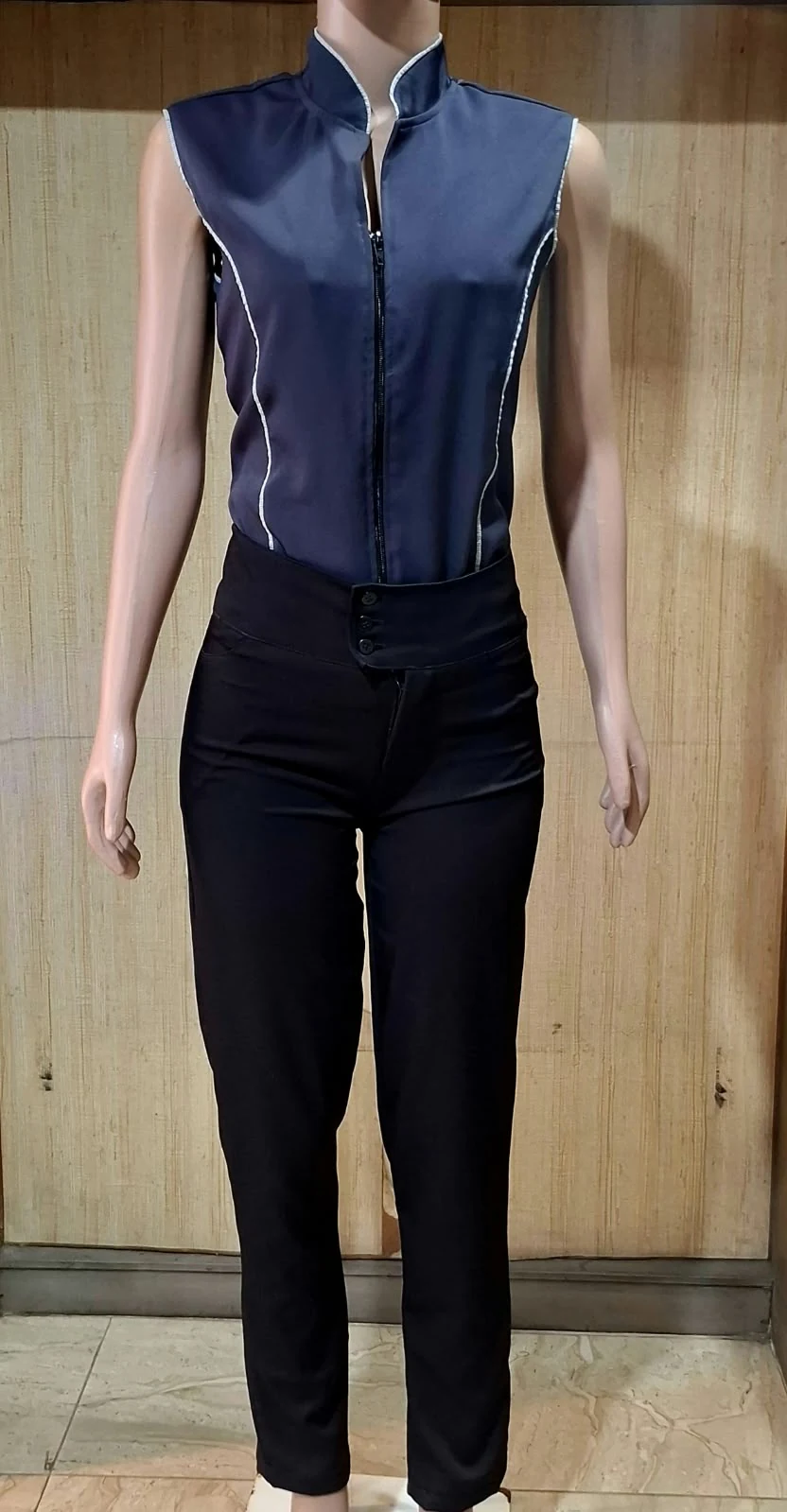 Manequim feminino com blusa azul-marinho sem mangas de gola alta, zíper preta e viés claro, e calça preta de cintura alta.