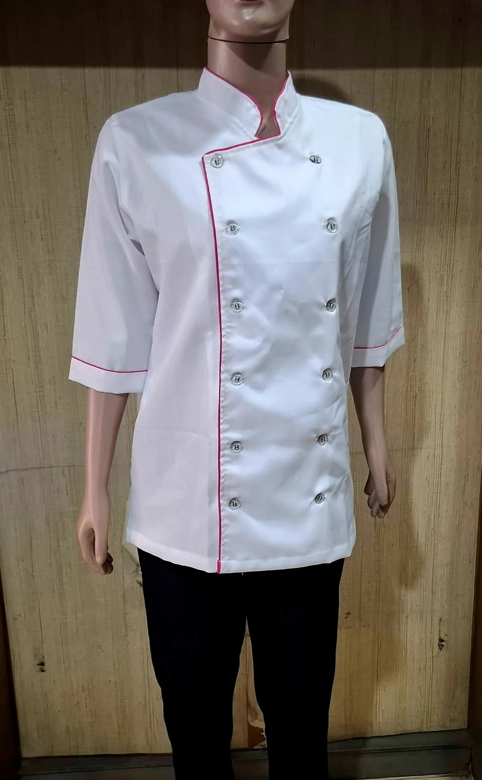 Manequim com jaqueta de chef branca de botões duplos, gola alta e viés rosa na gola e mangas, sobre calça preta.