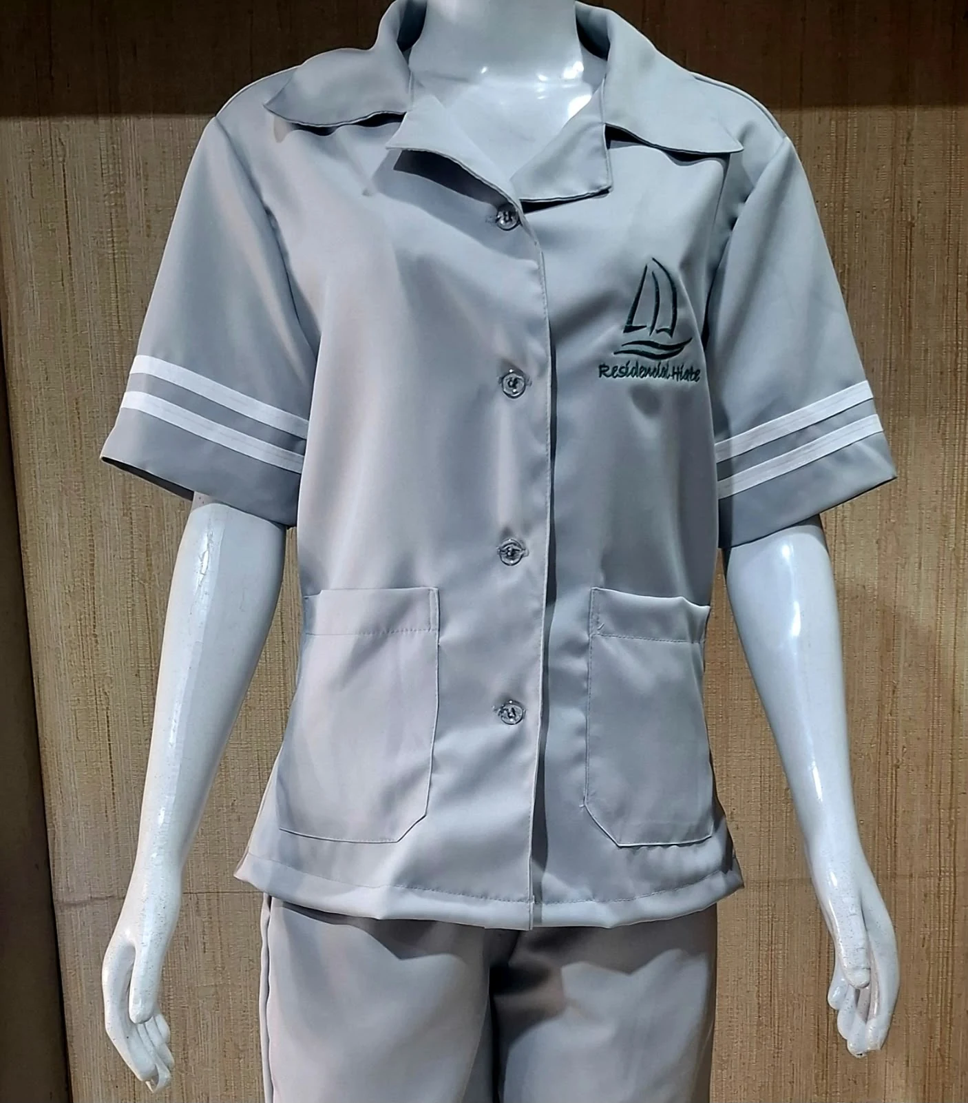 Manequim com camisa cinza de manga curta, listras brancas nas mangas, dois bolsos e bordado Residencial Hobbe no peito, com calça cinza.