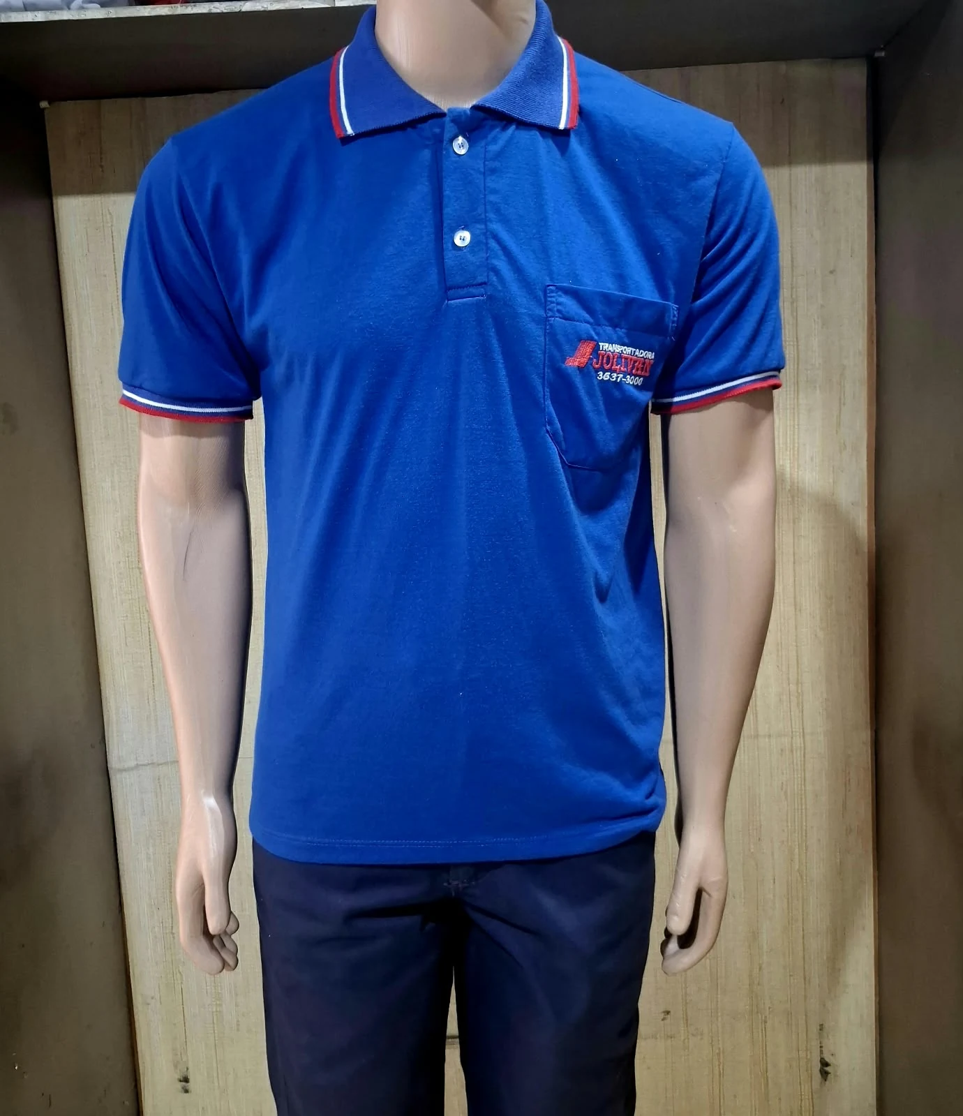 Manequim masculino com camisa polo azul de gola com listras vermelhas e brancas, bolso no peito e logotipo Transportadora Jolivens bordado.