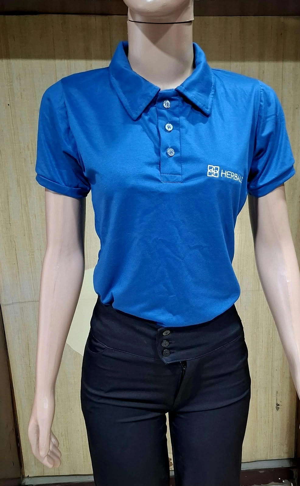 Manequim com camisa polo azul de manga curta, botões e logotipo branco no peito, e calça preta.
