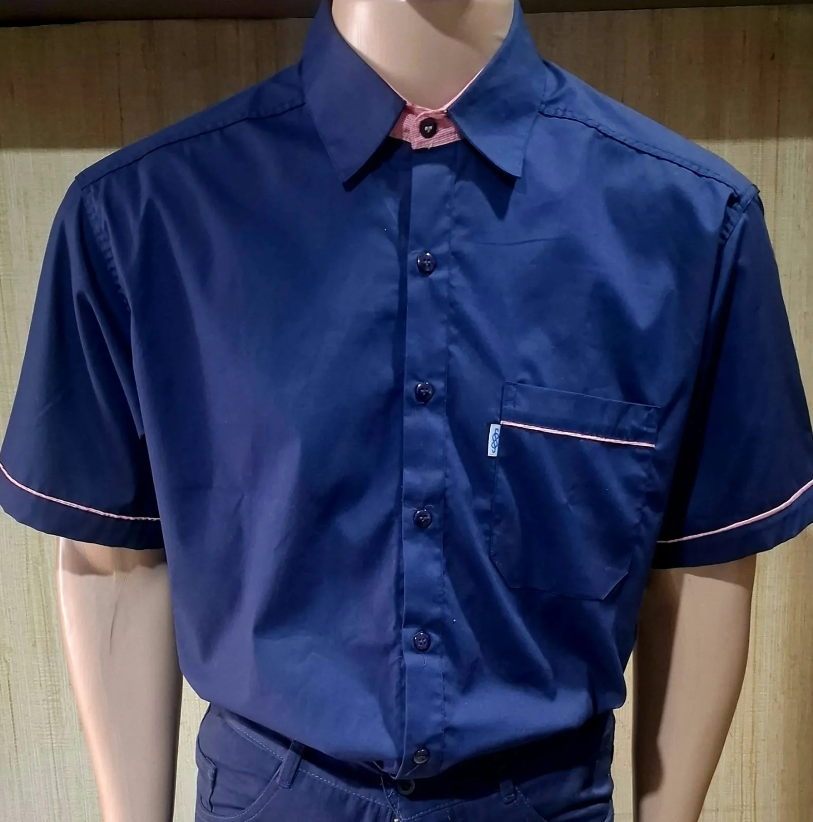 Manequim masculino com camisa azul-marinho de manga curta com viés rosa na gola, punhos e bolso.